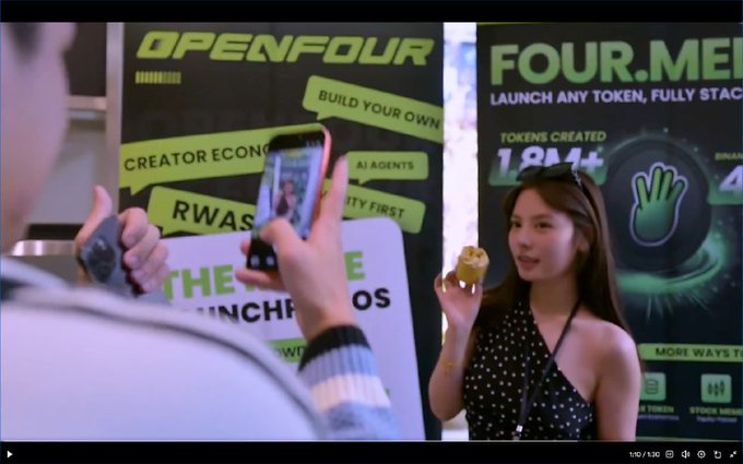 A woman stands at a Token2049 booth in front of banners for 'OPENFOUR' and 'FOUR.MEME' (showing '1.8M+ tokens created') while a person films her with a smartphone.