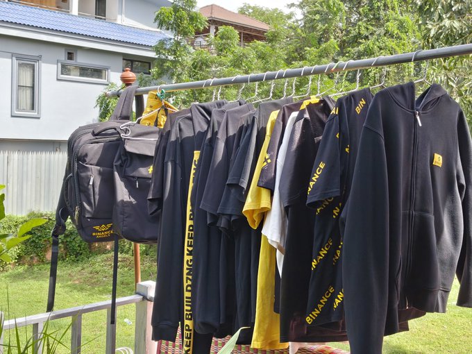 A clothesline outside a residential building displays several black Binance-branded hoodies and t-shirts hanging on hangers alongside a backpack.