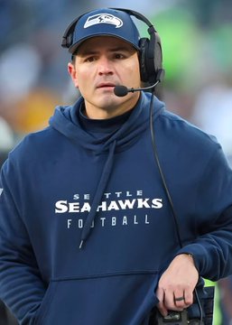 A Seattle Seahawks football coach wearing a navy "SEATTLE SEAHAWKS FOOTBALL" hoodie and headset stands on the sidelines, illustrating the post about head coach Mike Macdonald reflecting on his early leadership struggles with the team.