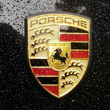 The Porsche crest emblem, featuring the iconic horse and antler design with "PORSCHE" and "STUTTGART" text, is shown in gold against a wet black background. The image relates to the news as it represents the luxury automaker ending its Web3 and NFT initiatives, including the Pioneer 911 NFT collection.