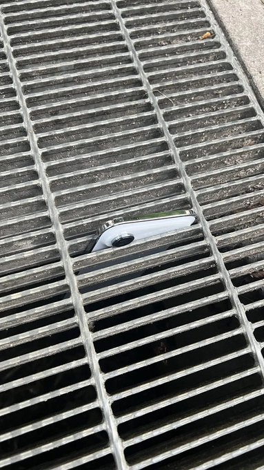 A smartphone lying face-down, partially fallen into the narrow gap between the metal slats of a storm drain grate.