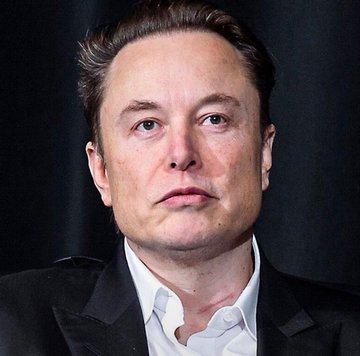 The image shows a close-up portrait of Elon Musk wearing a dark suit jacket and white collared shirt against a dark background, looking directly at the camera. The accompanying post relates to Musk's commitment to accelerating xAI's Colossus supercomputer project by living on-site in a motor home in Tennessee.
