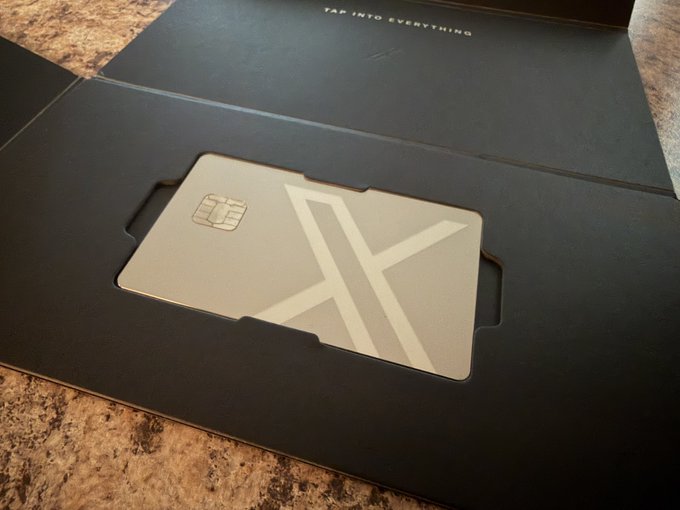 A sleek metallic card featuring a large "X" logo and an embedded chip rests in a black envelope, with "TAP INTO EVERYTHING" visible on the folder flap above, matching the X Money card being celebrated in the post.
