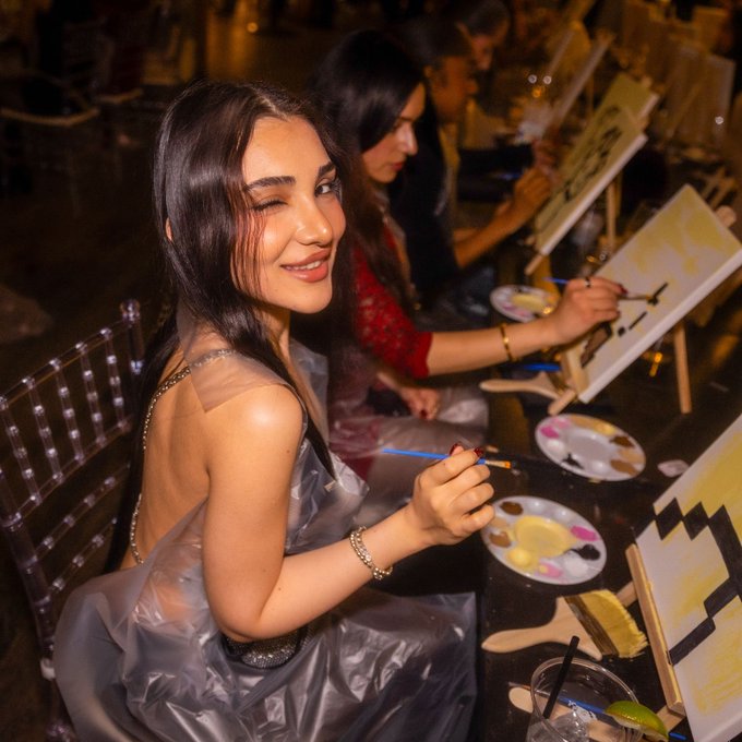 A young woman with long dark hair smiles at the camera while holding a paintbrush and palette at an art class, wearing a sheer grey dress over a dark top.