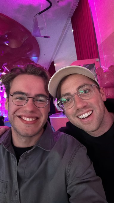 The image features two men wearing glasses, both smiling for a photo. They are standing close together with their arms around each other's shoulders, creating an intimate atmosphere. The background of the picture includes a pink wall that adds a pop of color to the scene. The caption of the image reads "So much love for the Zupateam @SuperteamDE Amazing summit & reunited with the people who got me into Solana!".
