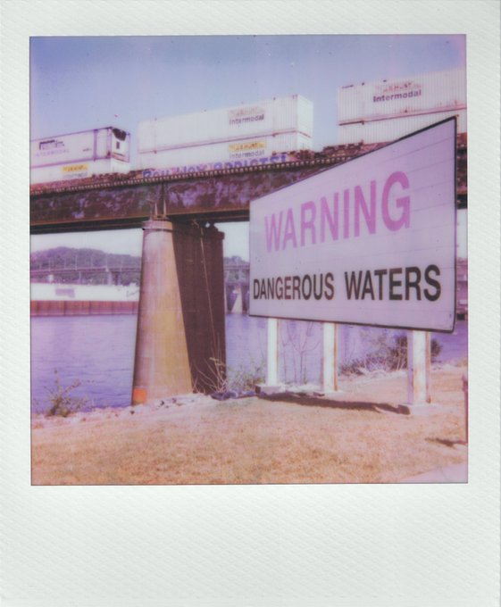 A Polaroid-style photo shows a railroad bridge over water with a sign reading 'WARNING DANGEROUS WATERS' and shipping containers labeled 'J.B. HUNT intermodal' passing overhead.