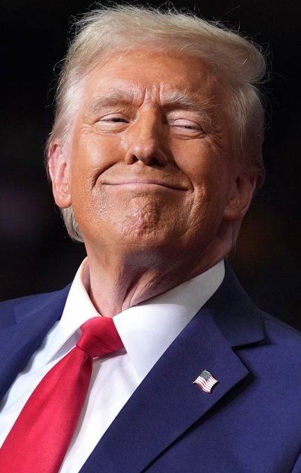 A close-up portrait of Donald Trump in a navy blue suit with a red tie and American flag lapel pin, smiling with his eyes closed against a dark background.