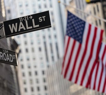 A "Wall St" and "Broad St" street sign sits beside an American flag in New York's financial district, with a skyscraper in the background—evoking the traditional home of US finance as the Depository Trust & Clearing Corporation (DTCC), which clears over $2 quadrillion in trades annually from those very blocks, moves toward blockchain-based tokenized securities.