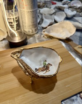 A decorative oyster shell-shaped dish with gold trim containing a small figurine of a cowboy on horseback, displayed on a bamboo surface with other shells and bottles in the background.
