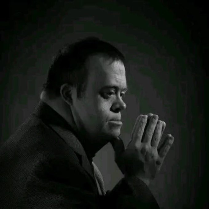A black and white profile photograph shows a man with his hands clasped together in a thoughtful or contemplative gesture against a dark background.