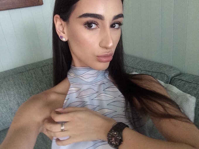 A selfie of a young woman with long dark hair wearing a light blue patterned halter top, showcasing a ring and a black watch.