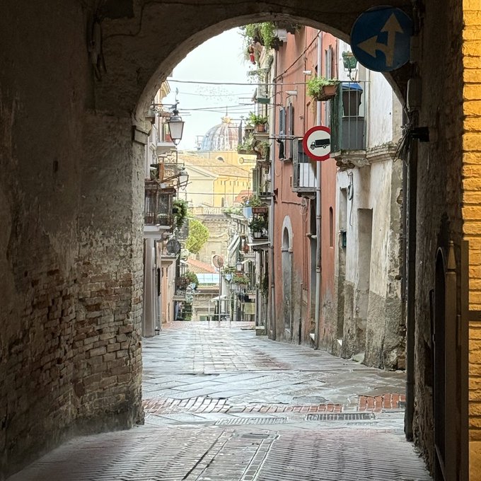 A narrow cobblestone street viewed through a brick archway in what appears to be an Italian city, with old buildings, balconies with plants, and traffic signs visible.