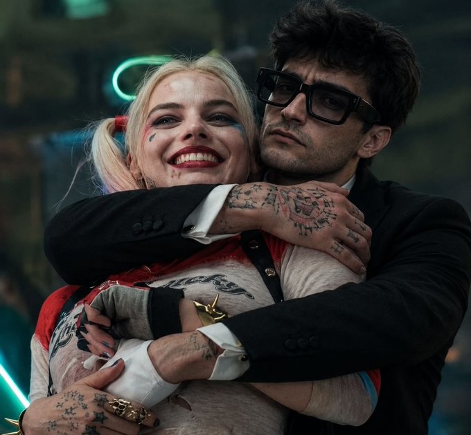 A smiling blonde woman with pigtails and face paint is being hugged from behind by a man in glasses, both wearing Harley Quinn and Joker-style costumes featuring visible "Daddy's Lil Monster" text and "Ha Ha Ha" tattoos on the man's hand, depicting the "Suicide Squad" aesthetic often used in cryptocurrency giveaway scams to lure victims into sharing wallet information.