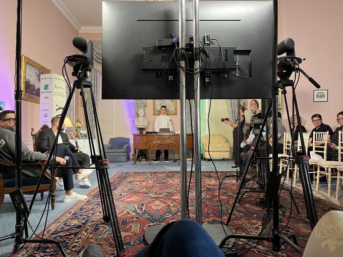 A view from behind a camera monitor showing a panel discussion being filmed in an ornate room, with a man seated at a desk and audience members visible.