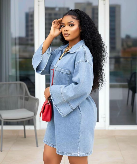 A woman with long curly black hair wearing an oversized denim jacket-dress and a gold chain necklace, holding a red handbag while standing on a balcony.