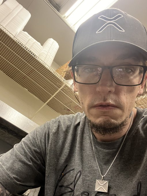 A man wearing a dark baseball cap with an 'X' logo, glasses, a gray t-shirt, and a silver pendant necklace takes a selfie inside what appears to be a food service area.