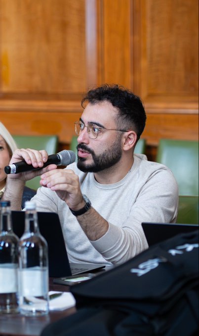 A bearded man wearing glasses and a white sweater speaks into a microphone while seated at a table with water bottles, gesturing with his hands.
