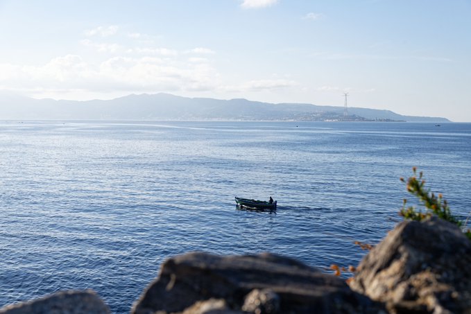 A scenic ocean view with a small boat in the water, distant mountains, and rocky outcroppings in the foreground.