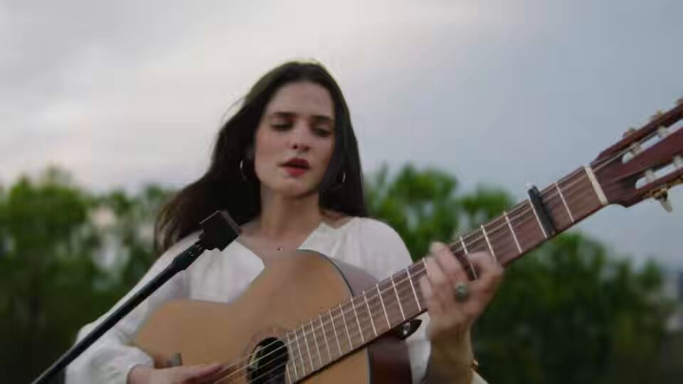 The image features a woman playing a guitar, with her hair blowing in the wind. She appears to be singing into a microphone while strumming the strings of her guitar. The photo captures this moment as she performs an acoustic song on stage. The background shows trees and a cloudy sky, creating a natural setting for the performance. The image also includes text that reads "POV: you wrote a song about what your final moment might feel like Blaze of Glory acoustic performance available on @manifoldxyz".