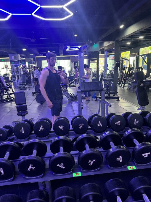 The image shows a man standing on a weight bench in a gym, holding onto some weights while taking care to keep his phone nearby. The man appears focused, possibly tracking a workout routine or monitoring progress. There are several black dumbbells lined up next to him on the ground, emphasizing his presence within the weight room environment. The focus of the image is on the man's activity in the gym and how it connects with the accompanying post about Bitcoin, personal health, and financial matters.