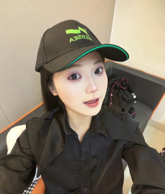 A selfie of a young woman wearing a black cap with a green Tesla logo and 'TESLA' text, dressed in a black shirt.
