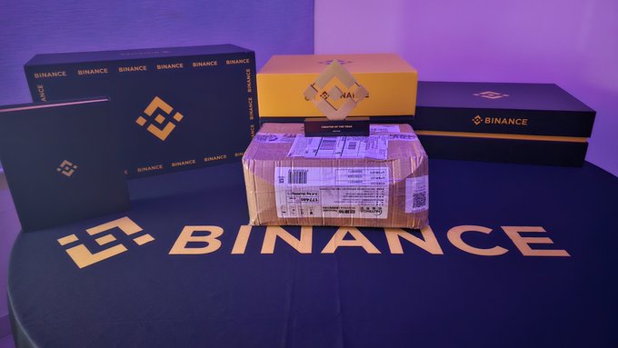 Display of Binance-branded merchandise including black boxes, a yellow box, and a shipping package, arranged on a black surface with the Binance logo.