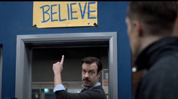 A scene with a yellow "BELIEVE" sign taped above a doorway, where a man with a mustache points upward while another person watches.