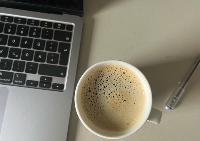 An overhead view of a workspace showing a laptop keyboard next to a cup of frothy coffee and a pen on a beige surface.