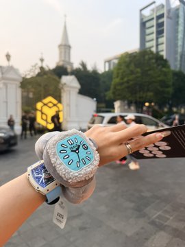 A first-person view of a hand wearing two watches—one with a blue strap and another with a furry gray strap—holding a phone, with a city street, church, and a yellow heart-shaped logo visible in the background.