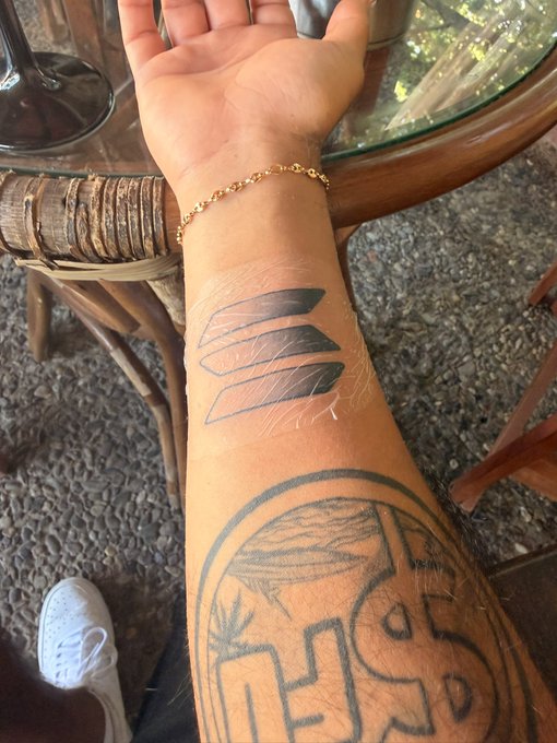 A close-up photo of a person's forearm displaying a fresh tattoo of three curved blades, with additional existing tattoos visible and a gold chain bracelet on the wrist.