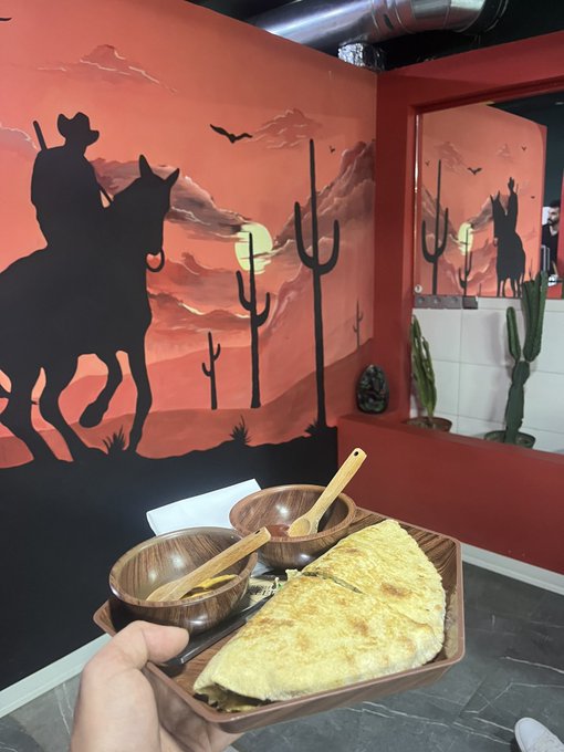 A person holds a wooden tray with a large quesadilla and small bowls of sauce in front of a vibrant mural depicting a cowboy on horseback, cacti, and a sunset desert scene, fitting the post's playful "Happy Tuesday, who wants some tacos?" message shared by @hellerincrypto.
