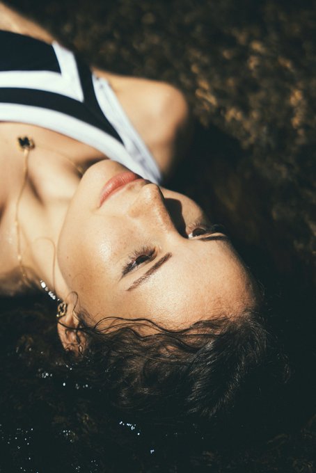 A close-up portrait of a woman with curly hair wearing a black and white striped top and a delicate necklace, lying down with her eyes closed.