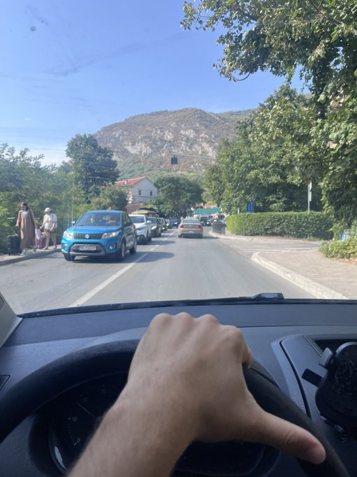 A first-person view from inside a car showing a hand on the steering wheel, driving on a road with a blue Suzuki parked on the left, mountains in the background, and pedestrians on the sidewalk.