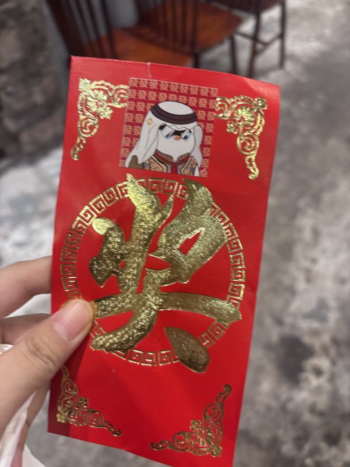 A hand holds a red Chinese New Year envelope featuring the gold Chinese character "發" (wealth/fortune) and a small image of a cartoon figure labeled "Binance Prince." The envelope ties to the caption's playful reference to "Binance Prince Um Hum" handing out festive red envelopes to bring good fortune.