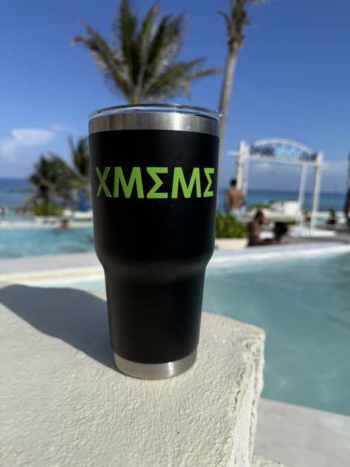 A black insulated tumbler displaying the green Greek letters 'ΧΜΣΜΣ' sitting on a ledge by a tropical resort pool with palm trees and the ocean.