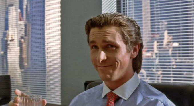A man in a blue shirt and red tie, resembling Patrick Bateman from American Psycho, smiles smugly while holding a glass in an office setting.