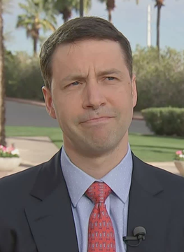A male reporter in a dark suit, light blue shirt, and red patterned tie delivers a news segment outdoors with palm trees visible in the background. His expression appears thoughtful, aligning with the accompanying post about Miller Value Partners criticizing MSCI's potential crypto treasury exclusions and their broader market implications.