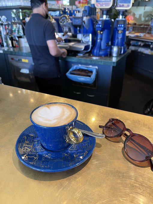 A blue ceramic cup of latte art sits on a matching saucer on a brass-colored bar counter, with sunglasses beside it and a bartender working in the background.