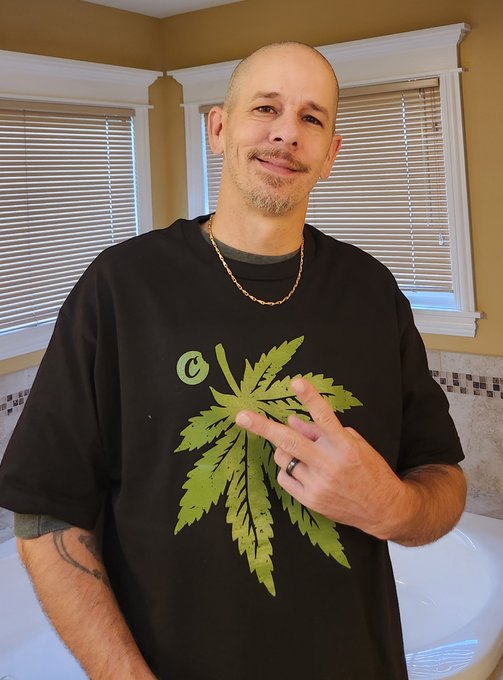 A bald man with a goatee stands in a bathroom wearing a black t-shirt featuring a large green cannabis leaf design with a 'C' logo, making a peace sign with his hand.
