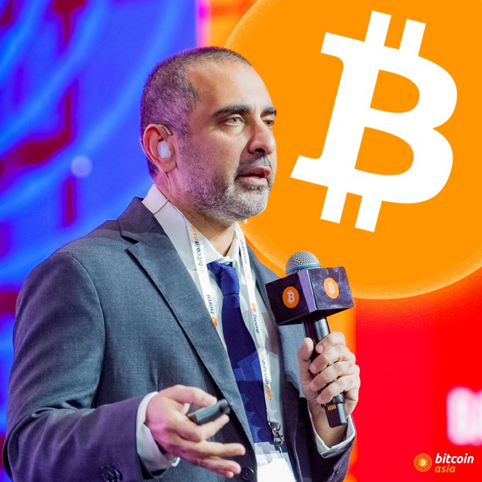 A man in a suit speaks into a Bitcoin-branded microphone at a conference, with a large orange Bitcoin logo displayed in the background and 'bitcoin asia' watermark visible.