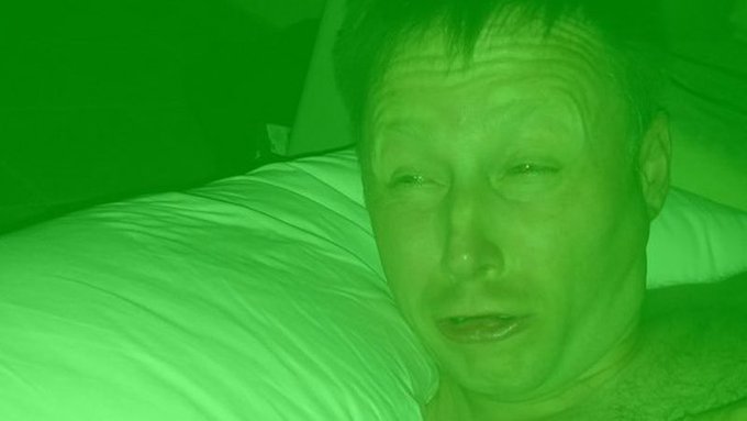 A green-tinted, low-light photograph shows a person with short hair looking toward the camera while resting near white pillows.