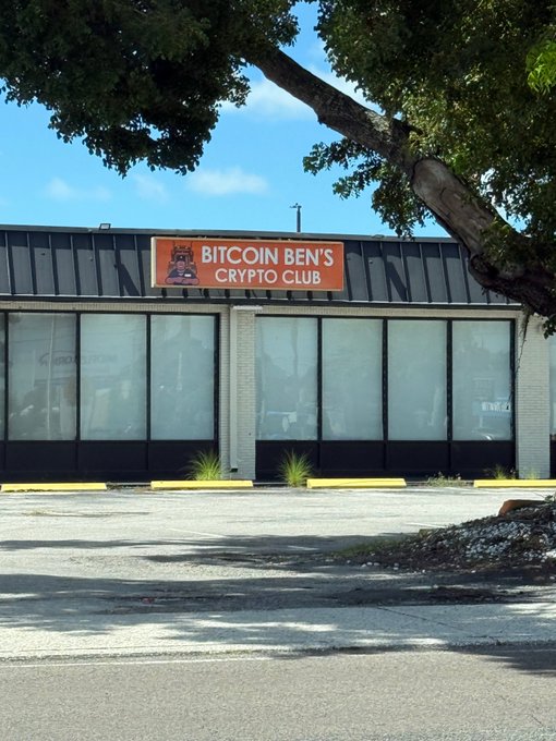 A storefront with a banner reading 'BITCOIN BEN'S CRYPTO CLUB' featuring a cartoon character logo, viewed from across a parking lot under a tree.