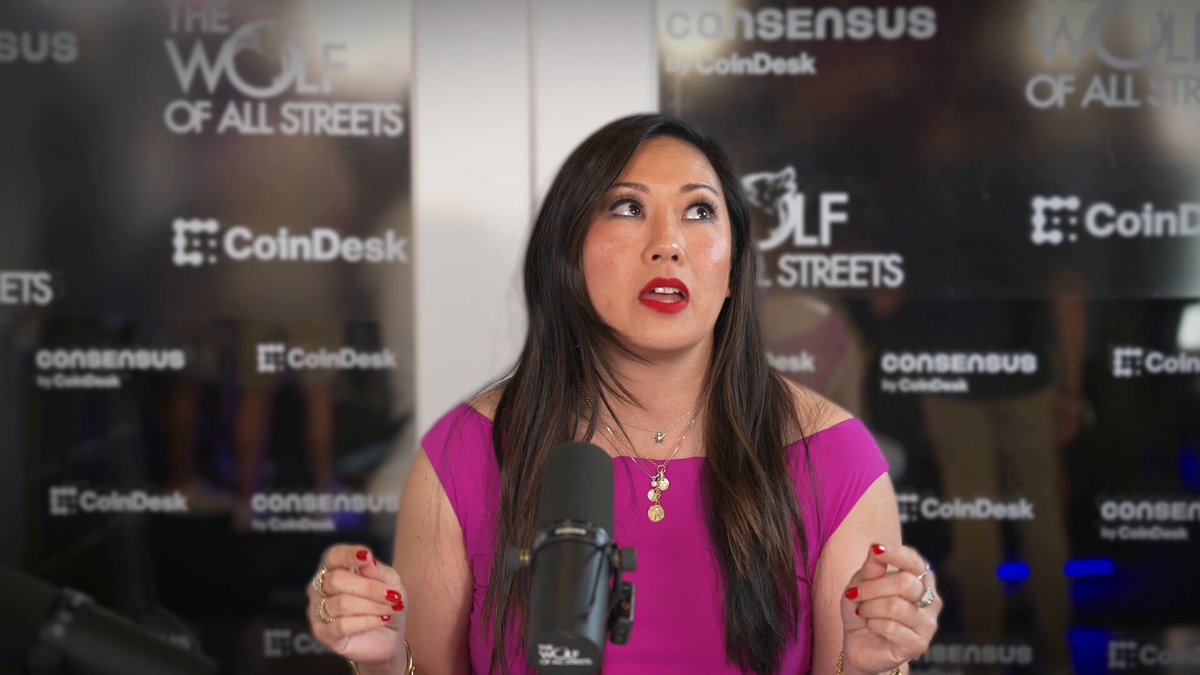 The image shows a woman wearing a pink dress standing at a microphone, giving a speech or presentation. The background features a black banner with white text that reads "The Wolf of All Streets" and "Consensusus CoinDesk". The caption below the image states "Let me put the scale of what'S happening into perspective, because it reframes how you should see $XRP", indicating an association between the woman's speech or presentation and a specific cryptocurrency.