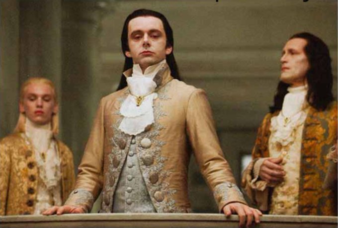 Scene from a period film showing a man in an elaborate 18th-century gold and cream embroidered coat with white cravat, standing on a balcony with other costumed figures.