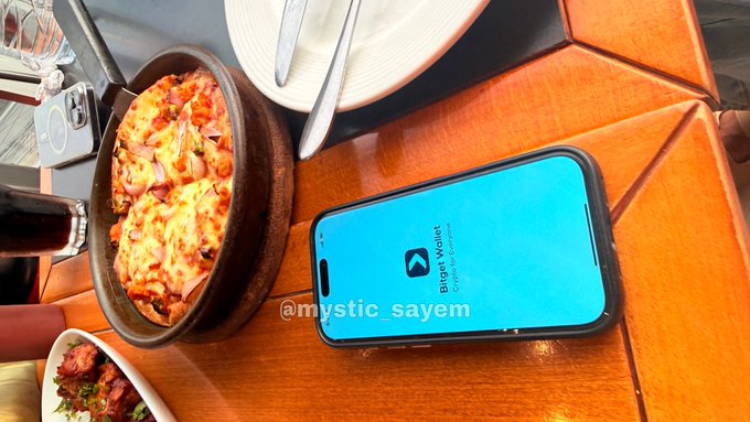 An overhead shot of a restaurant table showing a pan of baked food, white plates, and a smartphone displaying the 'Bitget Wallet Crypto & Bitcoin' app logo with the watermark '@mystic_sayem'.