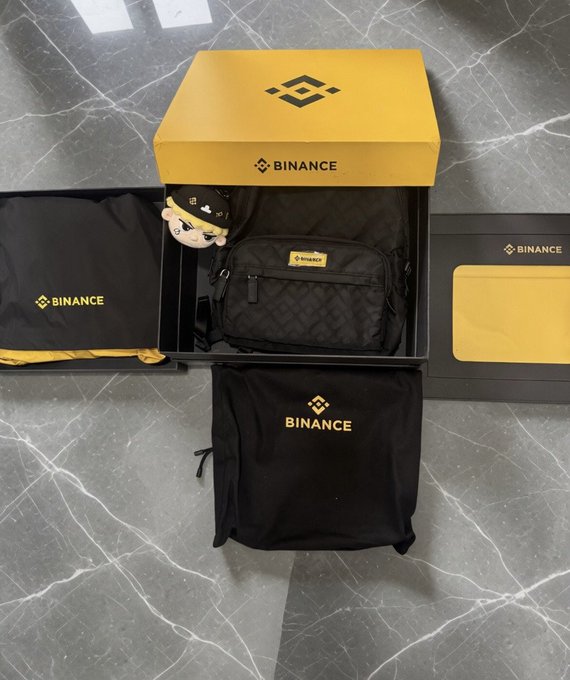 A black and yellow binance box, a black and white binance bag, and a black and gold binance shirt are displayed on a gray marble floor.