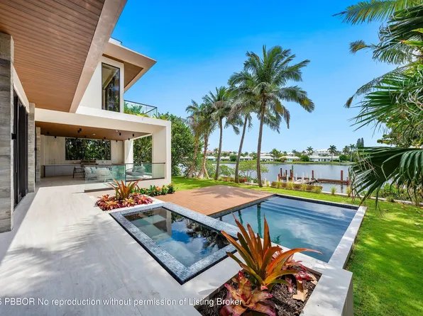 A modern waterfront home features a patio with a swimming pool, surrounded by palm trees and a lush green lawn.