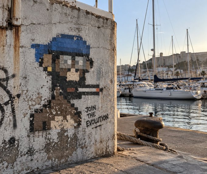 Pixel art street art on a concrete wall depicting a figure with sunglasses smoking, captioned 'JOIN THE EVOLUTION', set against a marina with sailboats and a fortified town in the background.