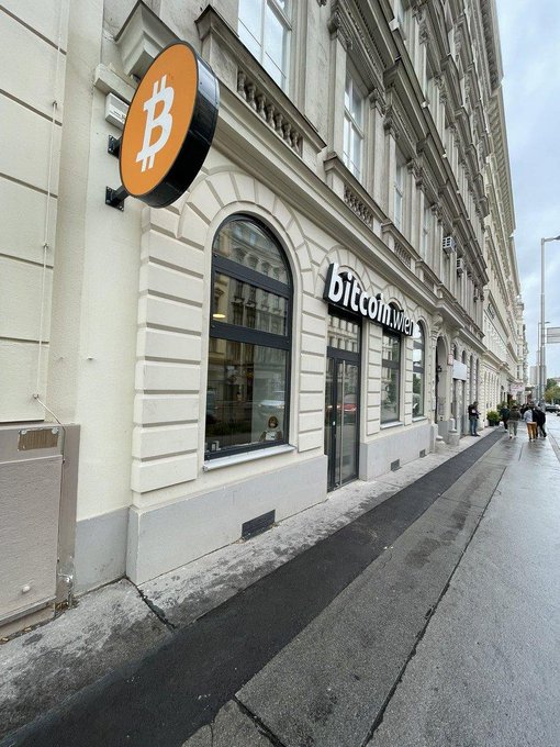 Exterior street view of a 'bitcoin.me' shop featuring an illuminated orange Bitcoin logo sign mounted on a classical-style building with arched windows.