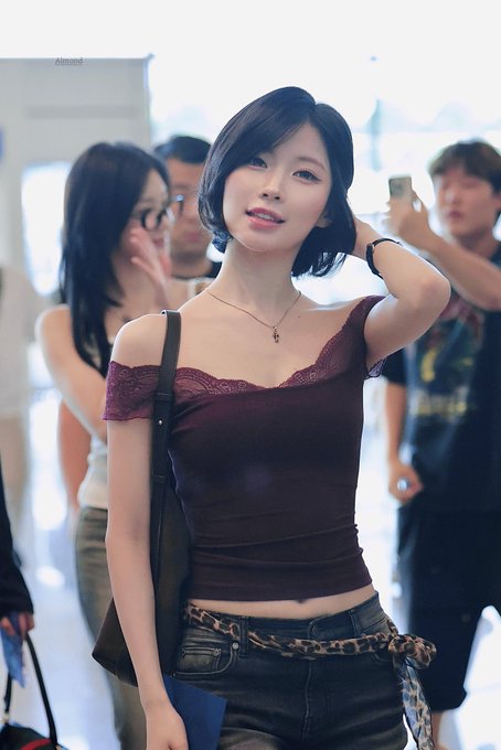 A young woman with short black hair wearing a burgundy off-shoulder top and jeans with a leopard-print belt stands in a crowd at what appears to be an airport, with the name 'Almond' faintly visible in the background.